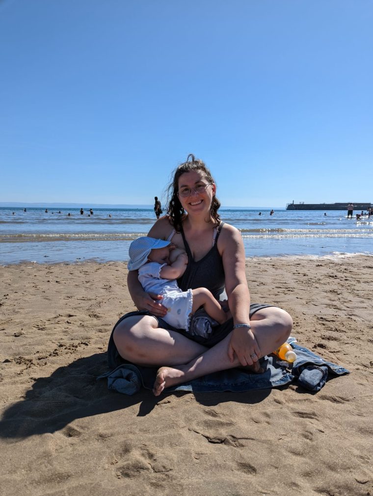 Image of a mum sitting cross-legged at the beach, breastfeeding her baby.